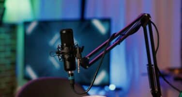 A podcast microphone mounted on an adjustable stand in a dimly lit recording studio with blue and purple ambient lighting.