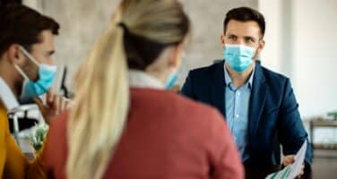 Three people wearing masks in a business meeting, sitting around a table with papers, in a modern office setting.