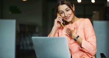 A woman in an orange blouse is happily talking on the phone while using a laptop in a modern office setting.
