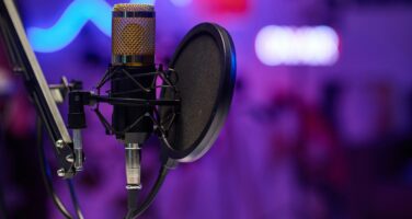 A microphone with a pop filter is mounted on a stand against a blurred background of purple and blue lights, suggesting a studio or recording setting.