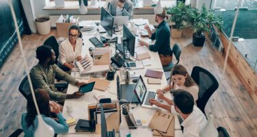 A diverse group of professionals collaborates in a modern office setting, seated around a large table filled with laptops, tablets, and documents. Green plants and natural light enhance the workspace's inviting atmosphere.