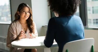 Two women are seated at a round table in a well-lit office space. One woman, with long hair, is actively engaging in conversation, while the other, with curly hair, listens attentively. They appear to be discussing something important.