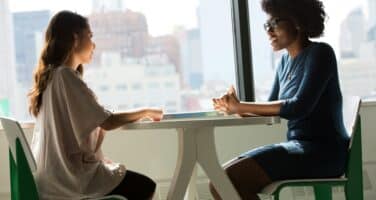 Two women are seated at a table, engaged in conversation. They are facing each other, with a city skyline visible through the window in the background. The woman on the left has long hair and wears a light top, while the woman on the right has curly hair and wears a blue dress.