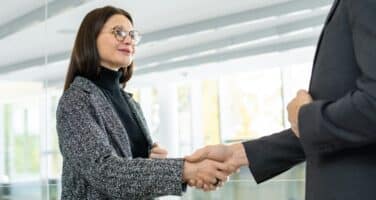 A woman with glasses and a gray coat shakes hands with a man in a suit in a modern office setting, symbolizing a professional meeting or agreement.