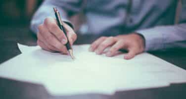 A hand holding a pen writes on a stack of papers on a table, suggesting an activity of signing or completing a document. A person in a light blue shirt is partially visible.