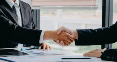 Two business professionals shaking hands across a desk, symbolizing a successful agreement or partnership. Documents and a laptop are visible on the table, indicating a professional setting.