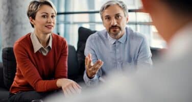 A man and woman sit on a couch, engaged in conversation with another person whose back is to the camera, in a bright, modern setting.