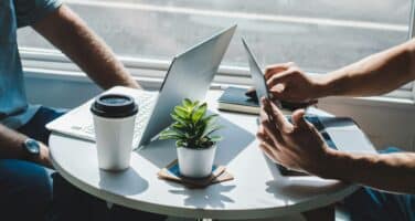 Two people sit at a small round table, working on laptops and a tablet. A coffee cup and a small potted plant are placed on the table, with sunlight illuminating the scene.