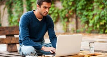 A young man sits outdoors at a wooden table, focused on his laptop. He wears a blue sweater and has a notebook and smartphone beside him, with greenery in the background.