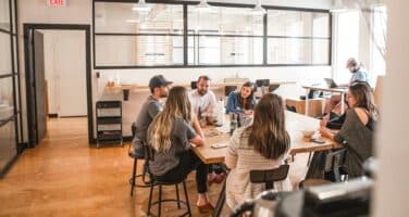 A group of eight people sits around a large table in a modern, brightly lit workspace. They are engaged in conversation, with a casual and collaborative atmosphere. Several individuals appear to be taking notes or discussing ideas, while a person in the background focuses on a laptop.