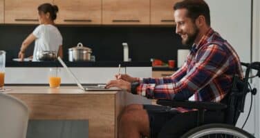 A man in a wheelchair works on a laptop at a modern kitchen table, while a woman cooks in the background. The scene conveys a sense of collaboration and comfort in a home environment.