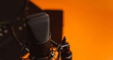 A professional microphone with a pop filter is set against an orange background, suggesting a recording or broadcasting environment.