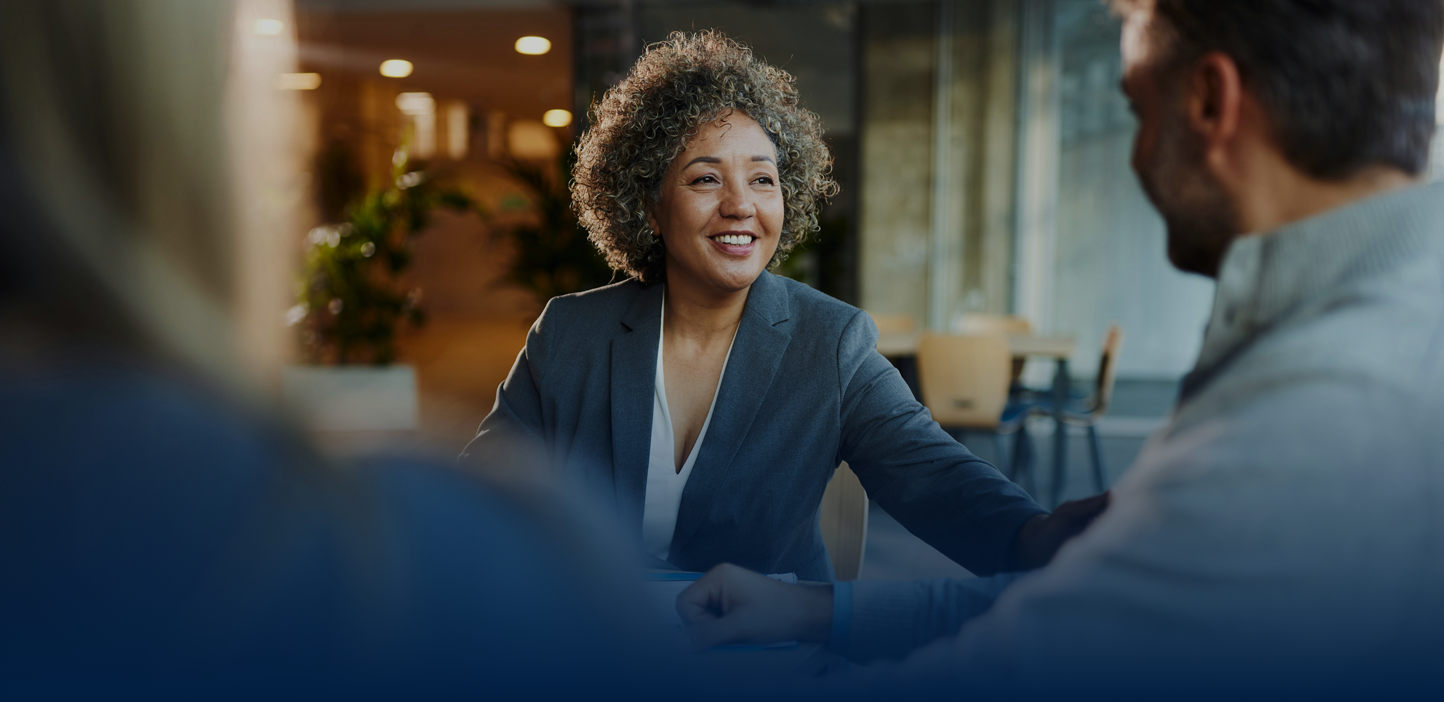 A woman with curly hair smiles while engaging in conversation with two individuals in a professional setting. The atmosphere is warm and inviting, highlighting a collaborative and friendly interaction.