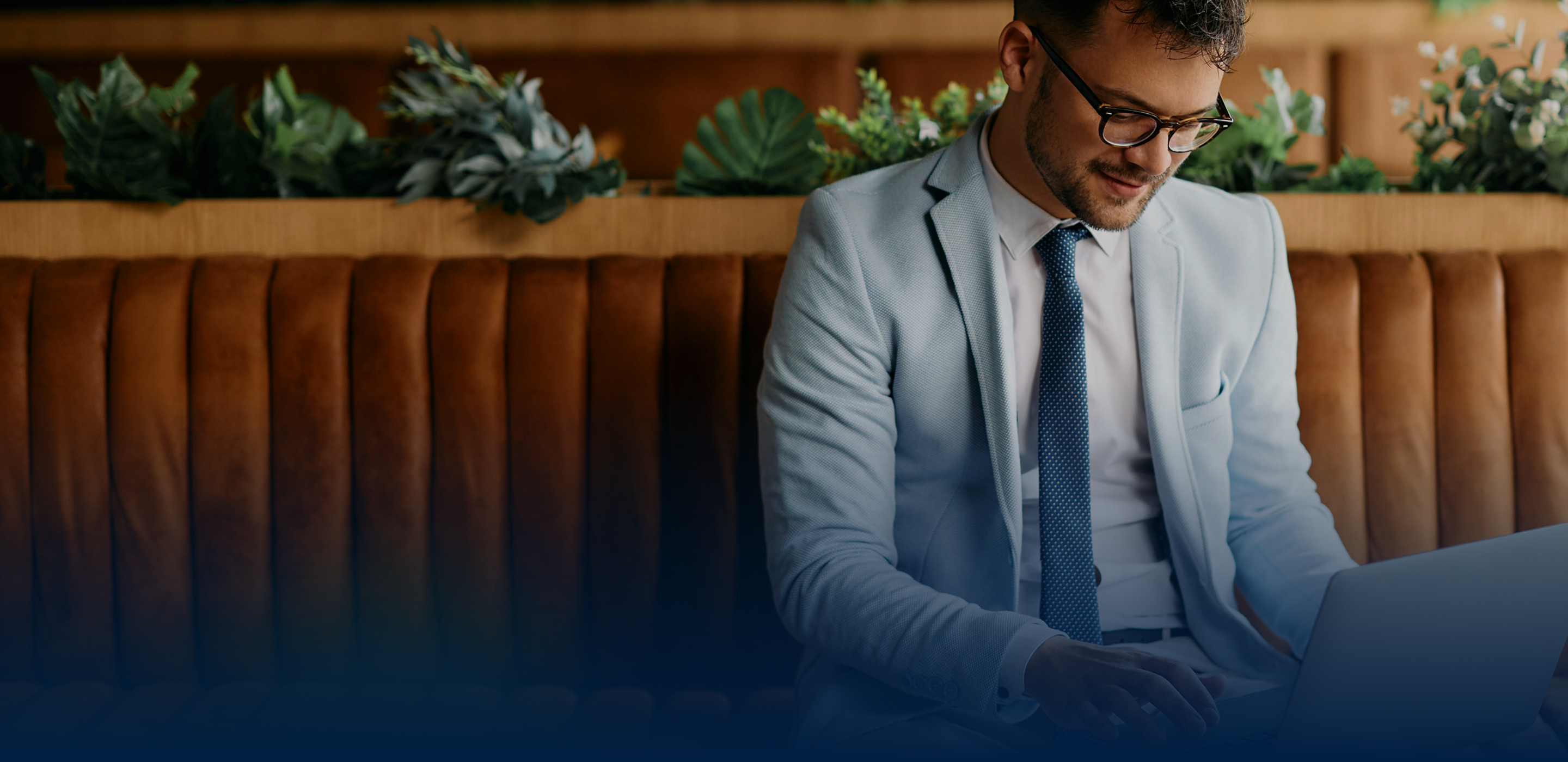 A man in a light blue suit and glasses sits at a table, smiling while working on a laptop. He is surrounded by green plants and a cozy, modern setting.
