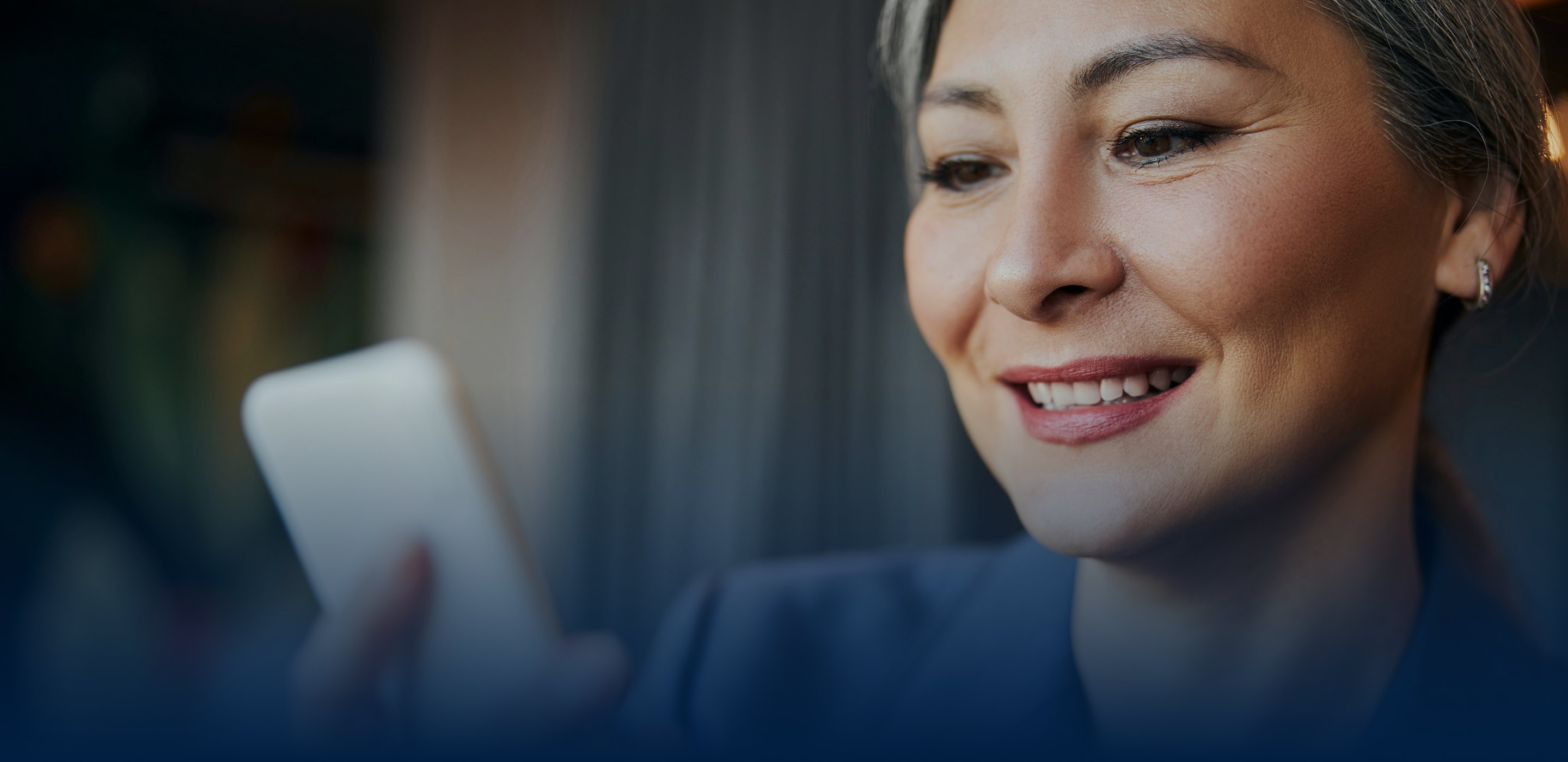 A smiling woman with gray hair holds a smartphone, looking at it with interest. The background is softly blurred, suggesting a cozy indoor setting.