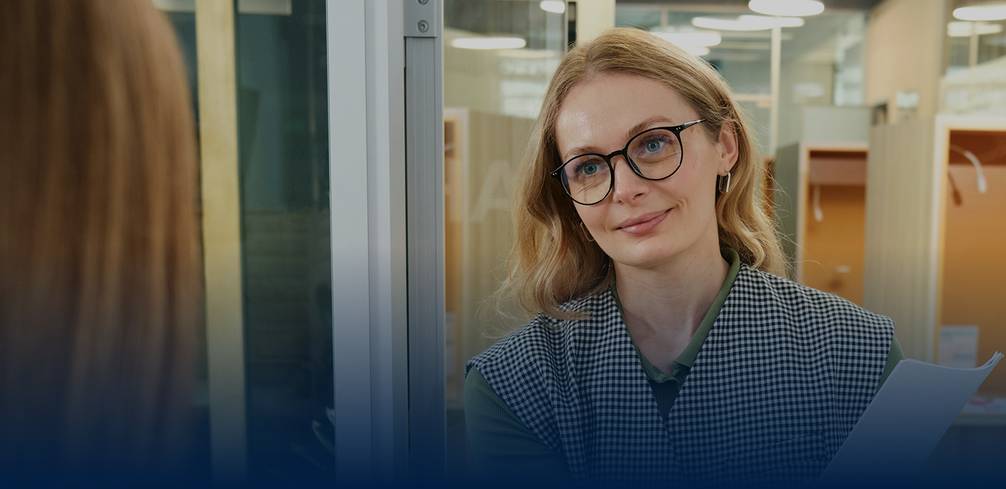 A woman with glasses and wavy hair smiles while engaged in conversation, standing in a modern office environment.