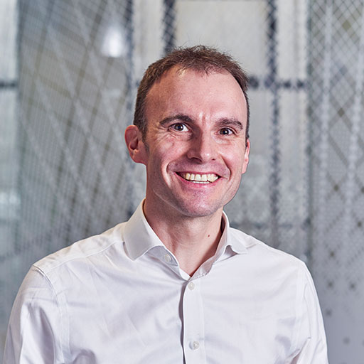 A smiling man in a white shirt stands in an office setting with a modern design, featuring blurred geometric patterns in the background.