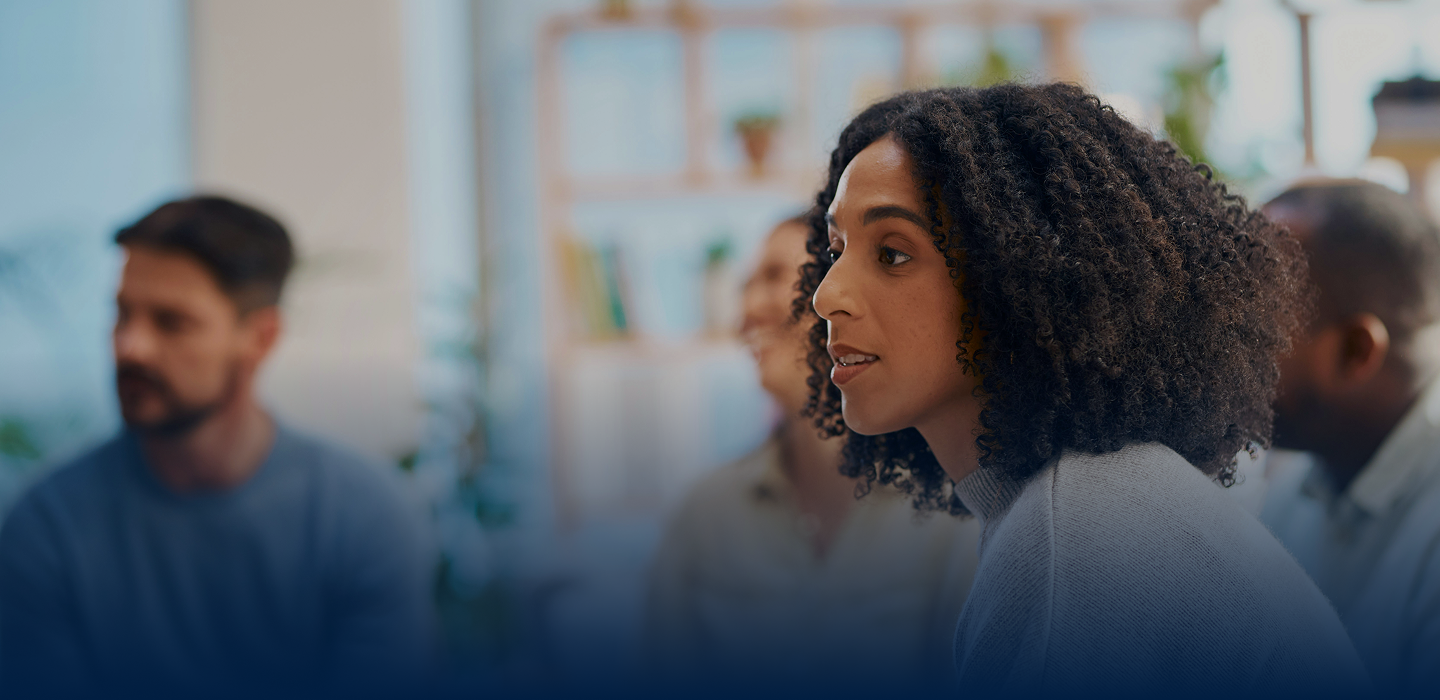 A group of diverse individuals engaged in conversation, with a focus on a woman with curly hair speaking intently. The setting appears casual and supportive, suitable for discussions or meetings.