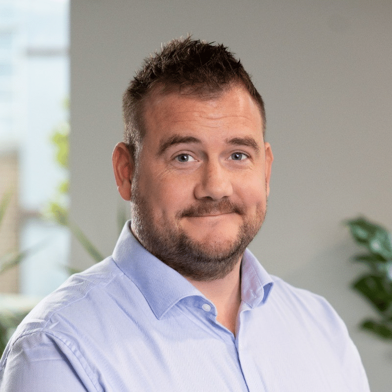 A man with short brown hair and a slight smile stands in an office setting, wearing a light blue shirt. Soft natural light filters in through a window behind him, and greenery is visible in the background.