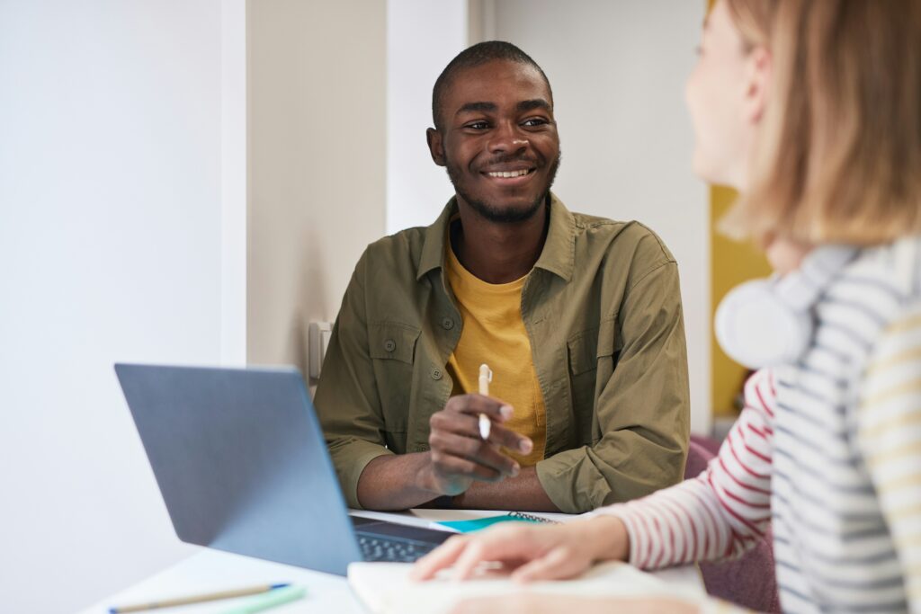A smiling young man engages in conversation with a woman, sitting at a table with a laptop open. The woman, who is partially visible, appears to be listening attentively. The setting suggests a casual study or collaboration environment.