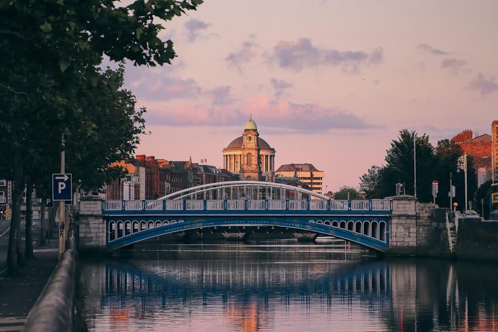 A blue bridge spans a calm river, with trees lining the bank. In the background, an elegant building rises against a pastel sky during sunset, reflecting the warm colors on the water's surface.