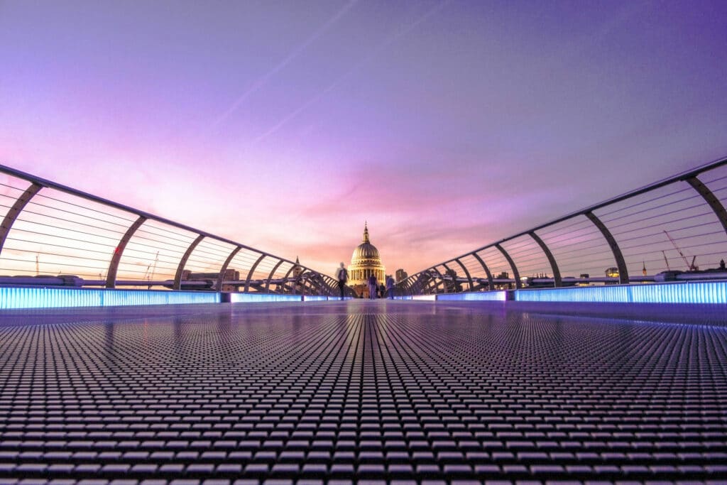 A perspective view of a modern bridge leading towards a historic dome structure, illuminated by soft purple and pink hues of dawn. The bridge’s grid pattern contrasts with the silhouette of people walking and the iconic architecture in the background.