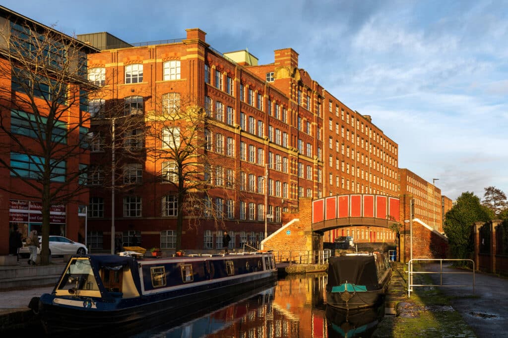 A sunlit canal scene featuring a historic red brick building with large windows, boats moored along the water, and trees lining the walkway. The bridge arching over the canal adds to the charming urban landscape.