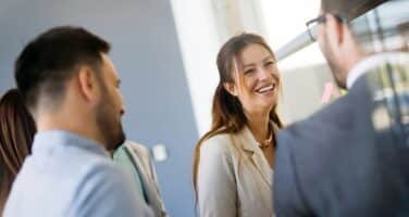 A group of professionals engage in a lively conversation, with one woman smiling as she speaks to two men, set in a bright, modern environment.