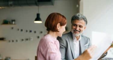 A smiling man and woman engage in a friendly conversation at a cafe, with the woman pointing to a document. The setting features modern decor and soft lighting, conveying a warm and inviting atmosphere.