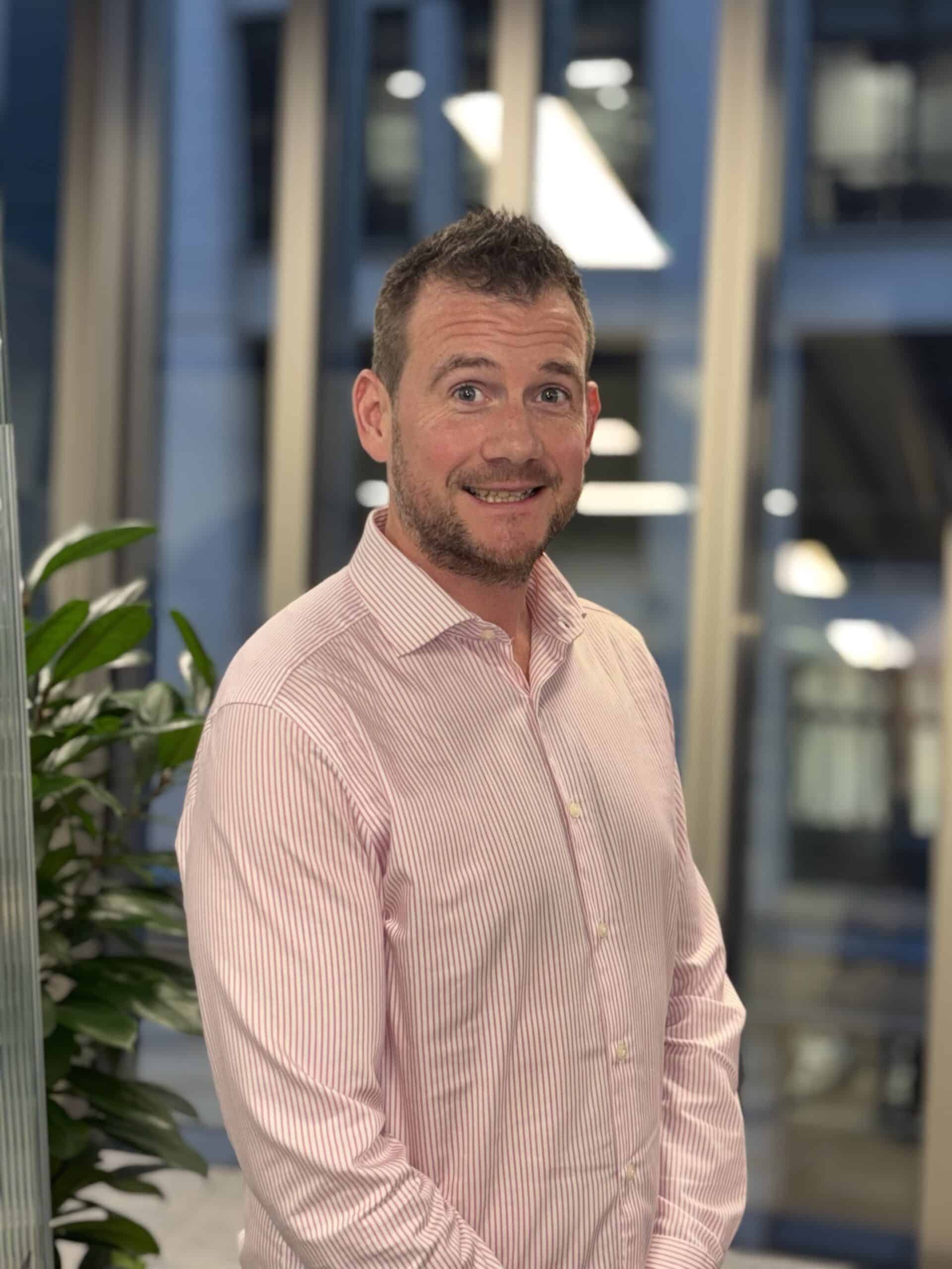 A man in a pink shirt stands in an office environment, smiling slightly with indoor plants and large windows in the background.