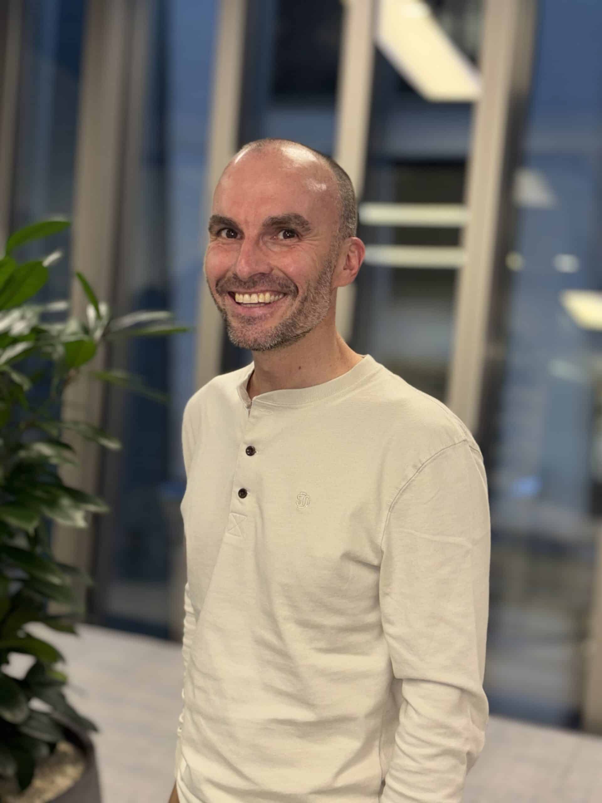 A man in a white shirt smiles while standing indoors, with modern office decor and plants in the background.