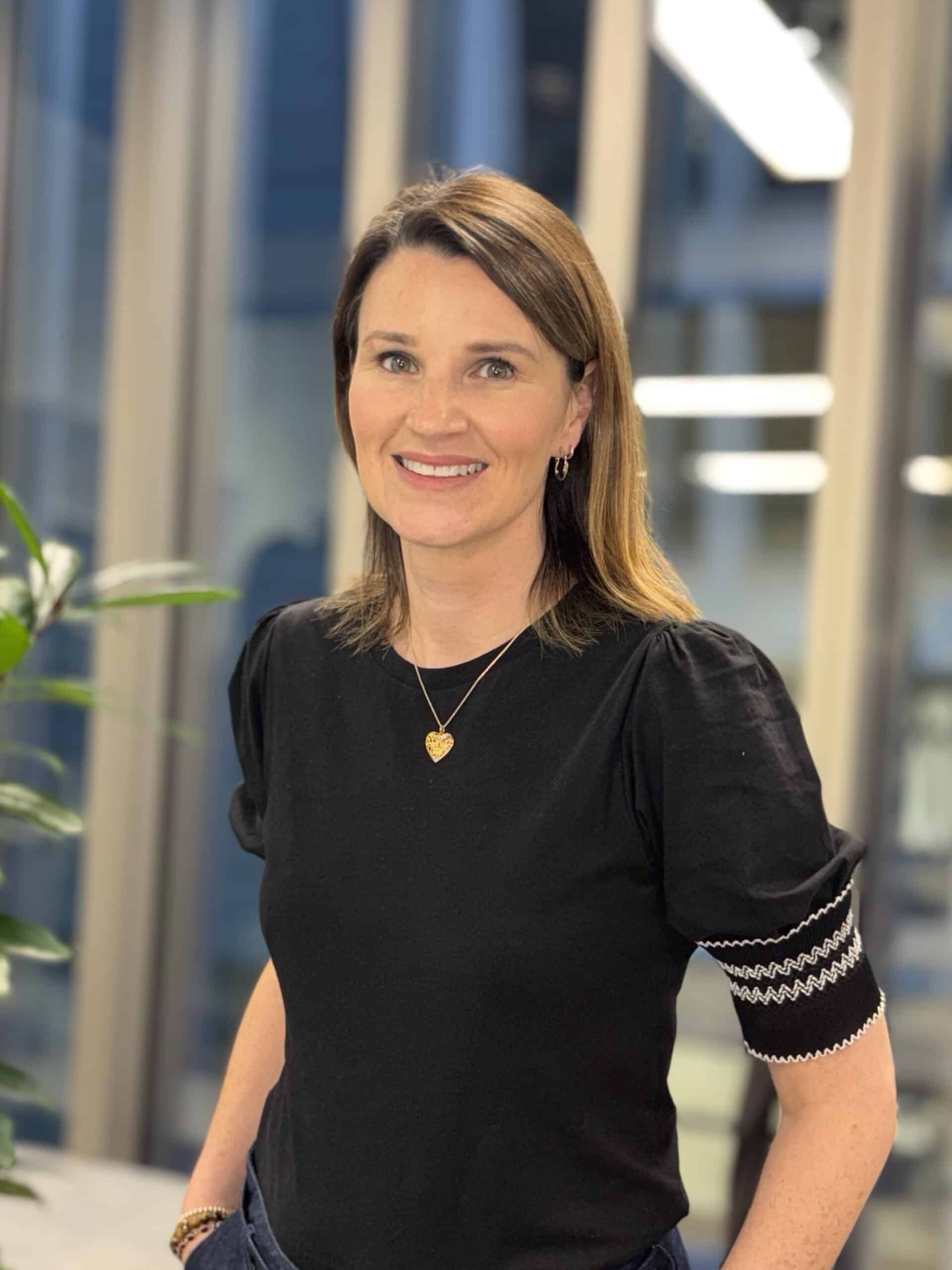 A smiling woman with brown hair wearing a black top stands indoors in front of a window.